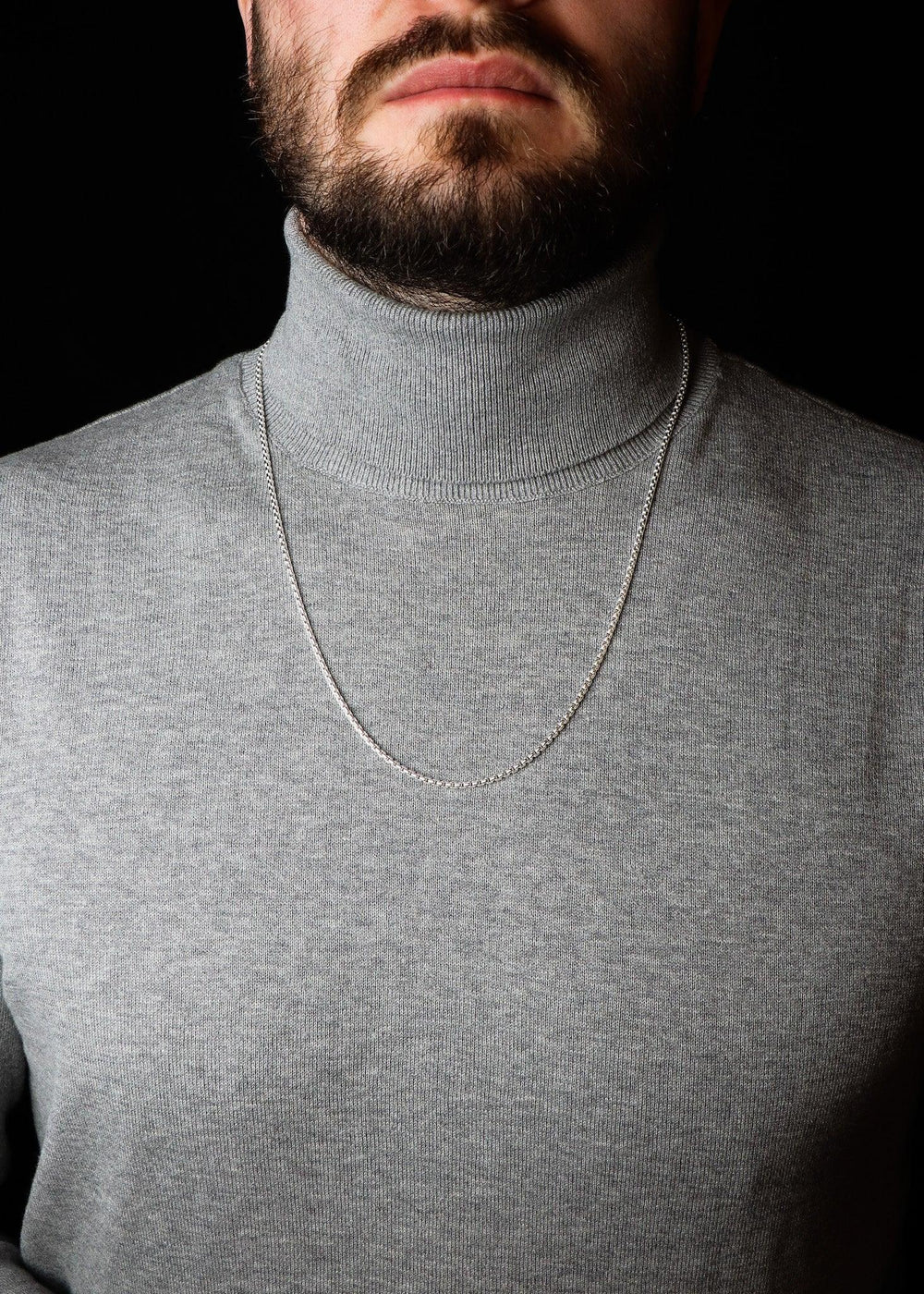 Man wearing a gray turtleneck sweater with a necklace against a black background