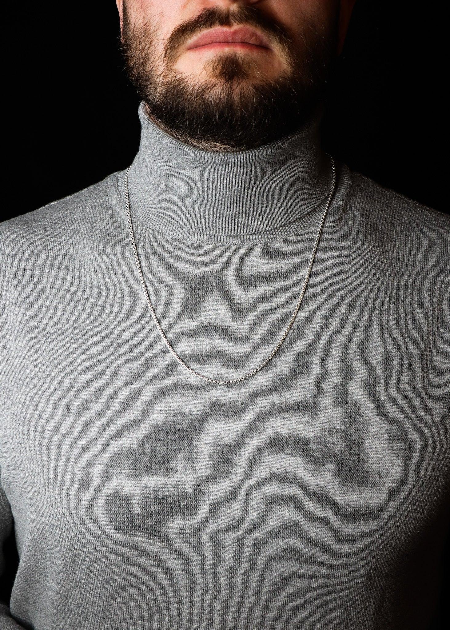 Man wearing a gray turtleneck sweater with a necklace against a black background